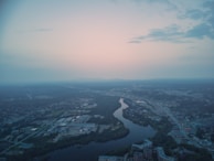 An aerial view of a cityscape at dusk. The scene is dominated by a winding river that cuts through the urban area, surrounded by a mix of residential, commercial, and industrial buildings. The sky is softly lit with pastel hues of pink and blue, indicating a setting or rising sun. Clouds are sparse, giving the sky a serene and expansive look. The city appears calm with little visible activity on the roads below.