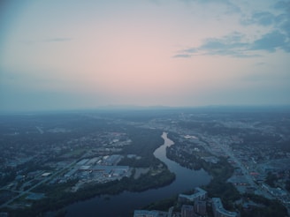 An aerial view of a cityscape at dusk. The scene is dominated by a winding river that cuts through the urban area, surrounded by a mix of residential, commercial, and industrial buildings. The sky is softly lit with pastel hues of pink and blue, indicating a setting or rising sun. Clouds are sparse, giving the sky a serene and expansive look. The city appears calm with little visible activity on the roads below.