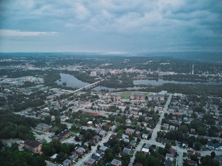 An aerial view of a cityscape featuring a blend of urban and natural elements. Buildings, roads, and residential areas are scattered throughout, surrounded by greenery and intersected by a river. The sky is overcast, suggesting an impending change in weather.