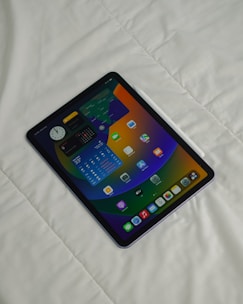 An electronic tablet lies on a bed with a white cover, displaying a colorful homescreen filled with app icons and widgets, such as a clock and a weather forecast. A white stylus is positioned on top of the tablet.