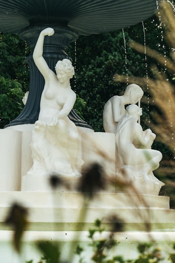 A classical marble fountain features intricately carved figures in various poses. The statues are set against a lush green backdrop, with delicate streams of water cascading from the edges of the fountain. The scene is partially framed by blurred foliage in the foreground.