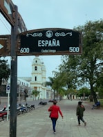 A street with a prominent sign reading 'España' and 'Ciudad Amiga' stands near the center. Two people walk along a wide, tree-lined sidewalk towards a tall, white bell tower. Several parked motorcycles line the street, and a person is seated on a bench under the trees.