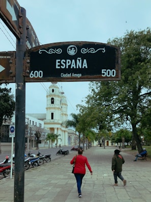 A street with a prominent sign reading 'España' and 'Ciudad Amiga' stands near the center. Two people walk along a wide, tree-lined sidewalk towards a tall, white bell tower. Several parked motorcycles line the street, and a person is seated on a bench under the trees.