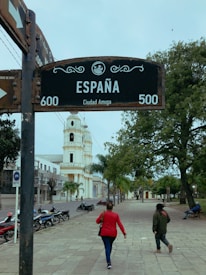A street with a prominent sign reading 'Espa&ntilde;a' and 'Ciudad Amiga' stands near the center. Two people walk along a wide, tree-lined sidewalk towards a tall, white bell tower. Several parked motorcycles line the street, and a person is seated on a bench under the trees.