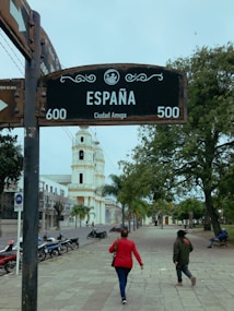 A street with a prominent sign reading 'Espa&ntilde;a' and 'Ciudad Amiga' stands near the center. Two people walk along a wide, tree-lined sidewalk towards a tall, white bell tower. Several parked motorcycles line the street, and a person is seated on a bench under the trees.