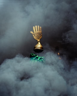 A golden hand-shaped sculpture with intricate writing is positioned above a metallic base, which is partially wrapped in a vibrant green cloth. The scene is enveloped in thick, swirling gray smoke, creating a mysterious and spiritual atmosphere.