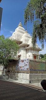 A large, white temple with multiple spires rises against a clear blue sky. The structure is surrounded by a metal fence adorned with posters and scripts. The foreground features a paved walkway and some trees, providing a tranquil atmosphere.