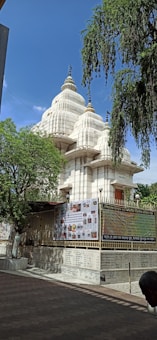 A large, white temple with multiple spires rises against a clear blue sky. The structure is surrounded by a metal fence adorned with posters and scripts. The foreground features a paved walkway and some trees, providing a tranquil atmosphere.