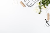 A white background with a variety of items arranged on the right side. There are white peonies with green leaves, a roll of twine, a pair of garden scissors, and a wire basket. The items are positioned to create a neat, organized composition with a minimalist aesthetic.