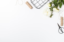 A white background with a variety of items arranged on the right side. There are white peonies with green leaves, a roll of twine, a pair of garden scissors, and a wire basket. The items are positioned to create a neat, organized composition with a minimalist aesthetic.