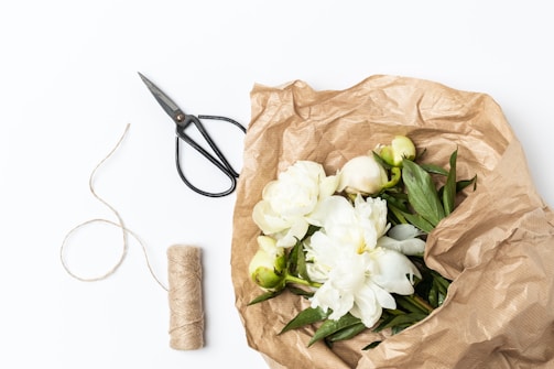 A bundle of white flowers with green leaves is wrapped in brown crinkled paper. Beside the wrapped flowers are a pair of black scissors and a roll of natural twine. The background is a clean white surface.