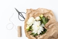 A bundle of white flowers with green leaves is wrapped in brown crinkled paper. Beside the wrapped flowers are a pair of black scissors and a roll of natural twine. The background is a clean white surface.