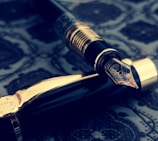 A luxurious fountain pen with intricate engravings on the nib, resting on a richly patterned surface. The pen's body features dark glossy material with gold accents, creating an elegant and vintage aesthetic.