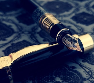 A luxurious fountain pen with intricate engravings on the nib, resting on a richly patterned surface. The pen's body features dark glossy material with gold accents, creating an elegant and vintage aesthetic.