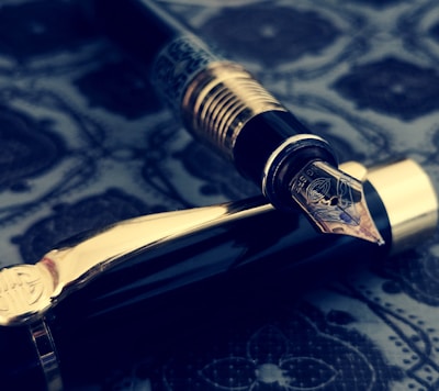 A luxurious fountain pen with intricate engravings on the nib, resting on a richly patterned surface. The pen's body features dark glossy material with gold accents, creating an elegant and vintage aesthetic.