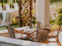 A cozy outdoor seating area features a round table surrounded by wicker chairs with white cushions. A glass vase with pink flowers is placed on a lace tablecloth. The setting is illuminated by warm, natural light and surrounded by lush greenery.