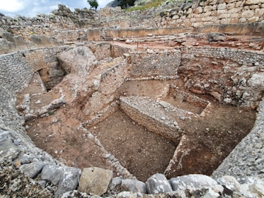 An ancient stone structure featuring a series of circular and rectangular formations. The stone walls are constructed using large, roughly hewn rocks, indicating historical architecture. The area appears to be an archaeological site, with layers of stone visible. The ground is dusty, and the sky is cloudy, adding a rustic atmosphere.