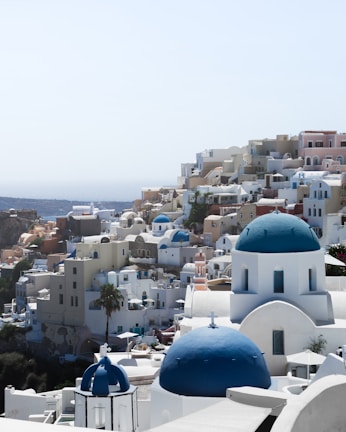 A picturesque coastal town with white buildings and striking blue domes, typical of Cycladic architecture. The town is built on a hillside with a view of the sea in the background. The buildings have a mix of arches and flat roofs, with narrow pathways between them.