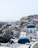 A picturesque coastal town with white buildings and striking blue domes, typical of Cycladic architecture. The town is built on a hillside with a view of the sea in the background. The buildings have a mix of arches and flat roofs, with narrow pathways between them.