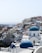A picturesque coastal town with white buildings and striking blue domes, typical of Cycladic architecture. The town is built on a hillside with a view of the sea in the background. The buildings have a mix of arches and flat roofs, with narrow pathways between them.