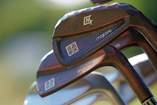 Close-up of several golf clubs with a unique hammered design, featuring inscriptions and branding including Japanese characters. The clubs are positioned at an angle, showcasing their metallic textures and craftsmanship.