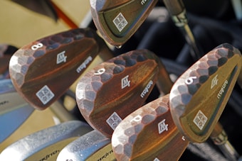 A close-up view of several golf clubs with wooden handles, featuring a distinct hammered texture. Each club is marked with numbers and logos, arranged neatly in a bag. The surface of the clubs appears polished with visible QR codes near the base.