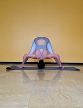 A person is performing a yoga pose on a gray exercise mat against a yellow wall. The pose involves bending forward with legs spread wide apart, hands touching the mat, and head hanging down towards the floor. The person is wearing a gray sports bra and gray leggings with light blue accents.