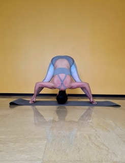 A person is performing a yoga pose on a gray exercise mat against a yellow wall. The pose involves bending forward with legs spread wide apart, hands touching the mat, and head hanging down towards the floor. The person is wearing a gray sports bra and gray leggings with light blue accents.