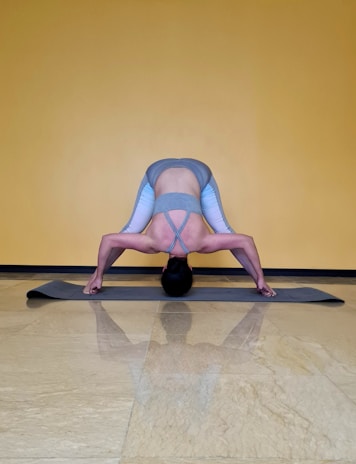A person is performing a yoga pose on a gray exercise mat against a yellow wall. The pose involves bending forward with legs spread wide apart, hands touching the mat, and head hanging down towards the floor. The person is wearing a gray sports bra and gray leggings with light blue accents.