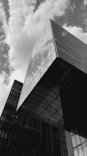 A striking black and white photograph of modern architecture featuring two tall, rectangular buildings with reflective glass surfaces. The angle provides a dynamic perspective with one building prominently towering upwards, capturing the reflections of the clouds and sky above.