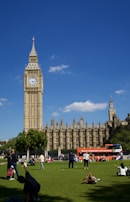A sunny day featuring the iconic clock tower and the Houses of Parliament. People are relaxing and engaging in various activities on the grassy area in the foreground. A bright red double-decker tour bus is parked nearby, and a few fluffy clouds are scattered in the clear blue sky.
