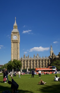 A sunny day featuring the iconic clock tower and the Houses of Parliament. People are relaxing and engaging in various activities on the grassy area in the foreground. A bright red double-decker tour bus is parked nearby, and a few fluffy clouds are scattered in the clear blue sky.