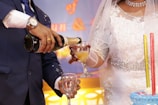 A close-up of a wedding scene featuring a bride in an elegant white dress with intricate beadwork and a groom in a dark suit. The groom is pouring champagne into a glass held by the bride. Nearby is a multi-tiered cake with tall decorative candles. The background shows soft lighting in hues of blue and yellow.