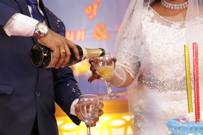 A close-up of a wedding scene featuring a bride in an elegant white dress with intricate beadwork and a groom in a dark suit. The groom is pouring champagne into a glass held by the bride. Nearby is a multi-tiered cake with tall decorative candles. The background shows soft lighting in hues of blue and yellow.