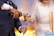 A close-up of a wedding scene featuring a bride in an elegant white dress with intricate beadwork and a groom in a dark suit. The groom is pouring champagne into a glass held by the bride. Nearby is a multi-tiered cake with tall decorative candles. The background shows soft lighting in hues of blue and yellow.
