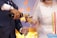 A close-up of a wedding scene featuring a bride in an elegant white dress with intricate beadwork and a groom in a dark suit. The groom is pouring champagne into a glass held by the bride. Nearby is a multi-tiered cake with tall decorative candles. The background shows soft lighting in hues of blue and yellow.