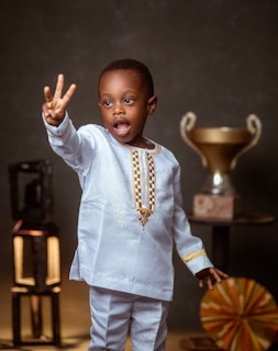 A young child wearing a traditional white outfit with gold embroidery gestures with three fingers raised. The background includes a large trophy on a stand and a decorative fan-like object.