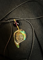A pendant with an artistic design depicting a face, featuring various colors such as green, brown, and gold. The pendant hangs from a black cord alongside a glossy black bead, set against a dark textured background.