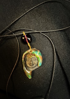 A pendant with an artistic design depicting a face, featuring various colors such as green, brown, and gold. The pendant hangs from a black cord alongside a glossy black bead, set against a dark textured background.
