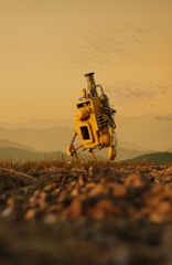 A solitary, yellow robot with a chimney-like structure on top stands on a rugged terrain. The landscape features a blurred foreground with grass and pebbles, and a hazy background of rolling hills under a pale orange sky.