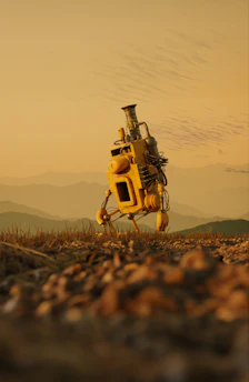A solitary, yellow robot with a chimney-like structure on top stands on a rugged terrain. The landscape features a blurred foreground with grass and pebbles, and a hazy background of rolling hills under a pale orange sky.