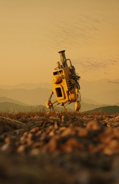 A solitary, yellow robot with a chimney-like structure on top stands on a rugged terrain. The landscape features a blurred foreground with grass and pebbles, and a hazy background of rolling hills under a pale orange sky.