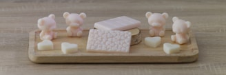 A wooden tray displays pastel-colored, bear-shaped and heart-shaped soaps, along with two rectangular, patterned soap bars. The scene is set on a wooden surface, giving a rustic and cozy feel.