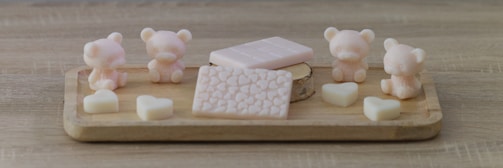 A wooden tray displays pastel-colored, bear-shaped and heart-shaped soaps, along with two rectangular, patterned soap bars. The scene is set on a wooden surface, giving a rustic and cozy feel.