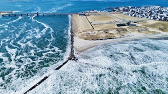 A coastal landscape featuring a clear blue ocean with waves crashing onto a sandy beach. A long pier extends into the water, leading to a populated area with buildings and infrastructure. The coastline is lined with rocky barriers and an expansive parking area is visible near the beach.