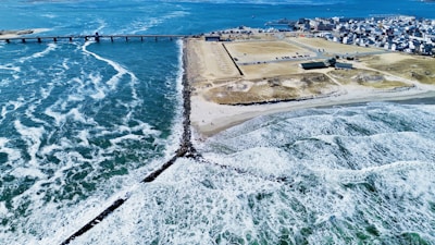 A coastal landscape featuring a clear blue ocean with waves crashing onto a sandy beach. A long pier extends into the water, leading to a populated area with buildings and infrastructure. The coastline is lined with rocky barriers and an expansive parking area is visible near the beach.