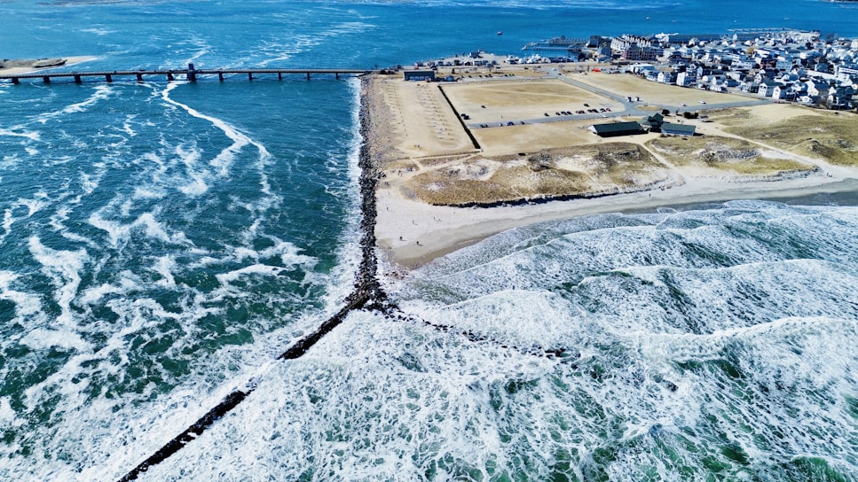 A coastal landscape featuring a clear blue ocean with waves crashing onto a sandy beach. A long pier extends into the water, leading to a populated area with buildings and infrastructure. The coastline is lined with rocky barriers and an expansive parking area is visible near the beach.