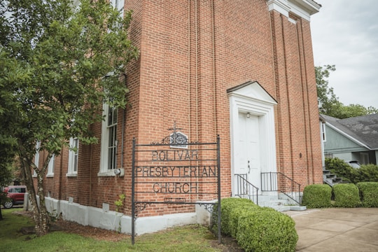A large, brick-built Presbyterian church with a white door and steps leading to the entrance. There is an ornate iron sign in front of the building indicating that it is the Bolivar Presbyterian Church, established in 1821. Trees and manicured bushes surround the church, with a gray building visible in the background.