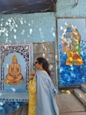 A person in a yellow outfit and denim jacket stands in front of a wall featuring two colorful paintings. One depicts a seated figure adorned with jewelry in a meditative pose. The other portrays a blue-skinned deity embracing a companion. Light filters through overhead, creating dappled patterns on the wall.