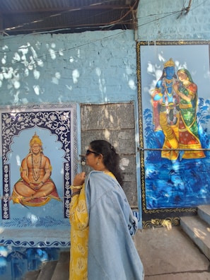 A person in a yellow outfit and denim jacket stands in front of a wall featuring two colorful paintings. One depicts a seated figure adorned with jewelry in a meditative pose. The other portrays a blue-skinned deity embracing a companion. Light filters through overhead, creating dappled patterns on the wall.